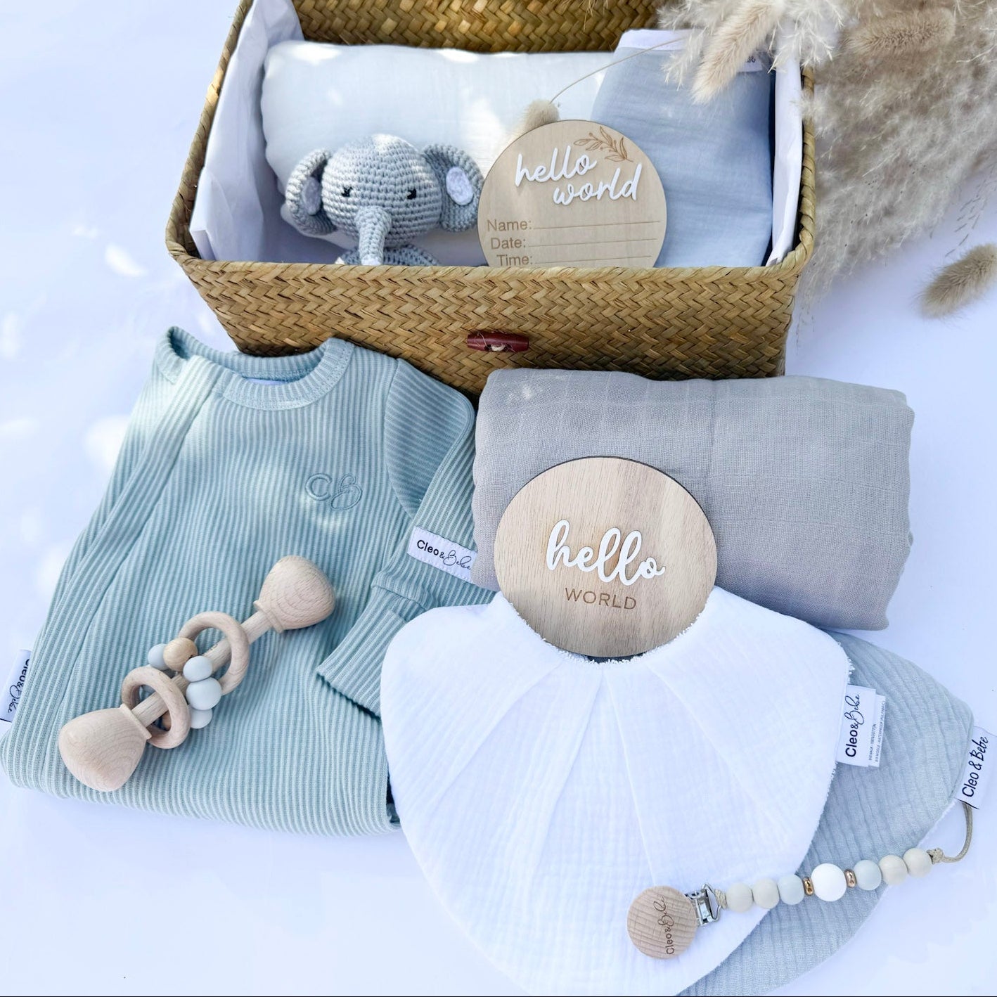 A gift box set with baby items including a blue muslin wrap, a gray classic bib, a waterproof bandana bib, a pacifier clip, a wooden rattle, and a blue onesie. The items are displayed neatly in a basket with a lid.