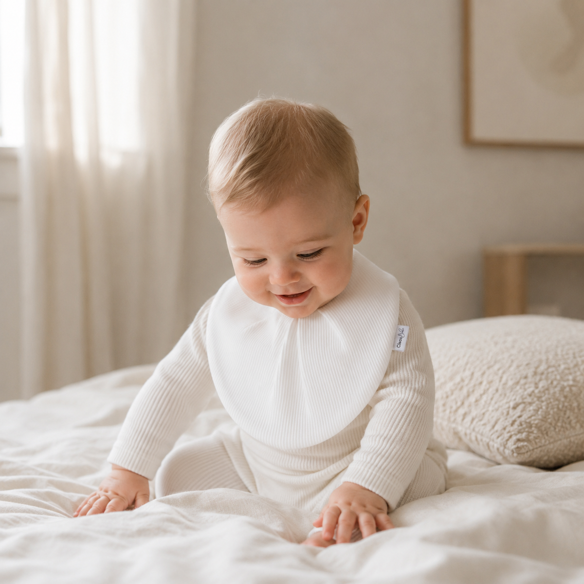 A baby wearing a white ribbed long-sleeve bodysuit and matching bib sits on a white bed in a softly lit bedroom.