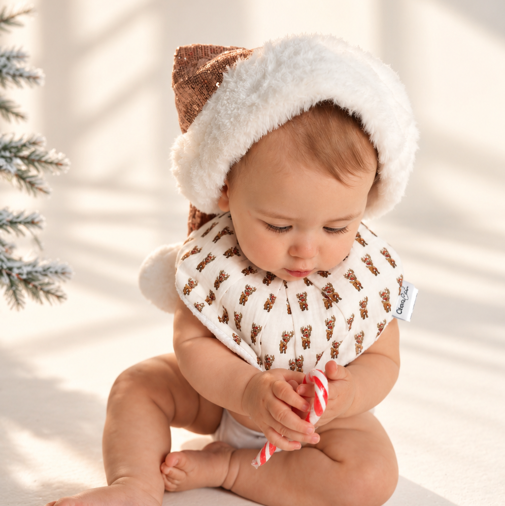 A baby sitting on a white floor wearing a white fuzzy Santa hat with brown ears and a white bib with a brown reindeer pattern, holding a red and white striped candy cane.