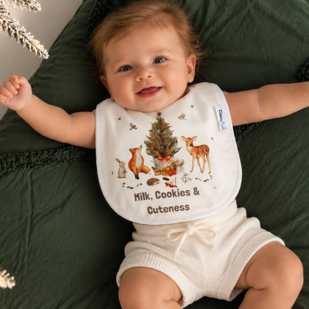 A baby lying on a dark green blanket wearing a white bib with a Christmas tree and deer illustration and the text 'Milk, Cookies & Cuteness'.