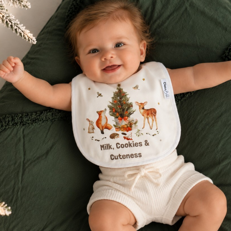 A baby lying on a dark green blanket wearing a white bib with a Christmas tree and deer illustration and the text 'Milk, Cookies & Cuteness'.