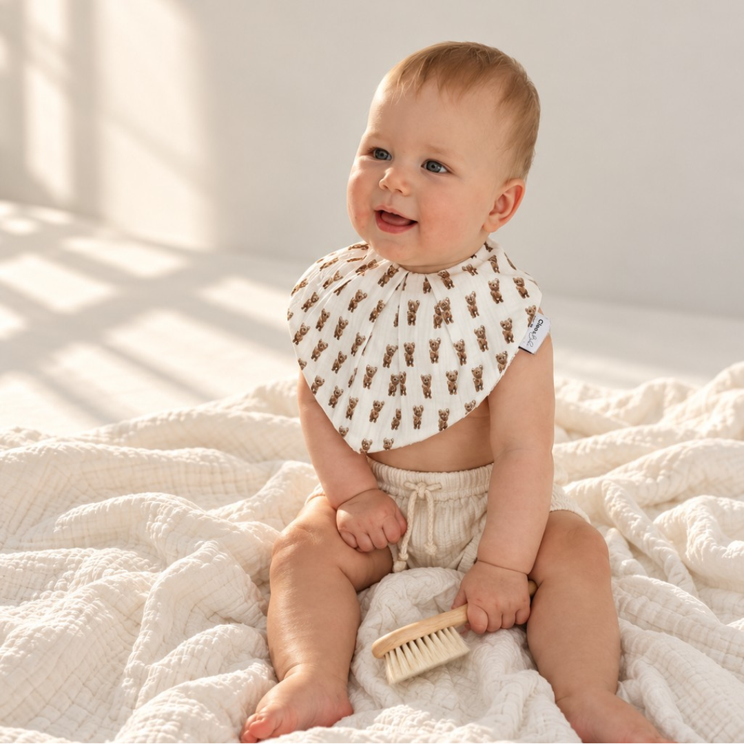 A baby sitting on a textured cream blanket in a brightly lit room, wearing a cream bib with a brown reindeer pattern and holding a small wooden brush.