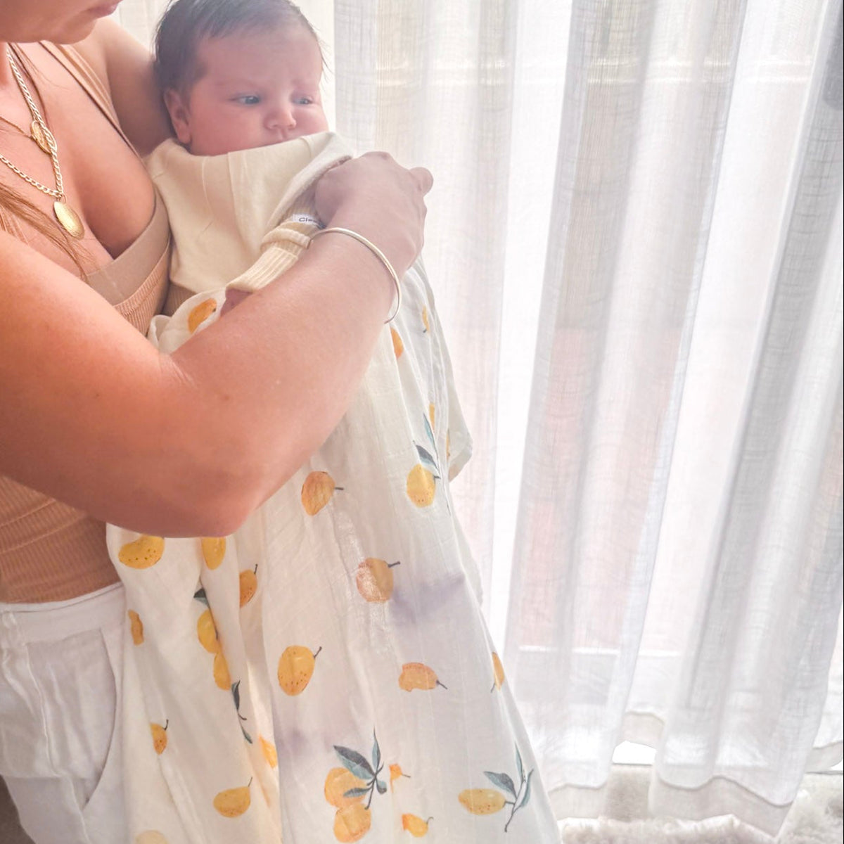 Woman holding a baby wrapped in a yellow and white blanket with a lemon pattern, standing in a softly lit room.