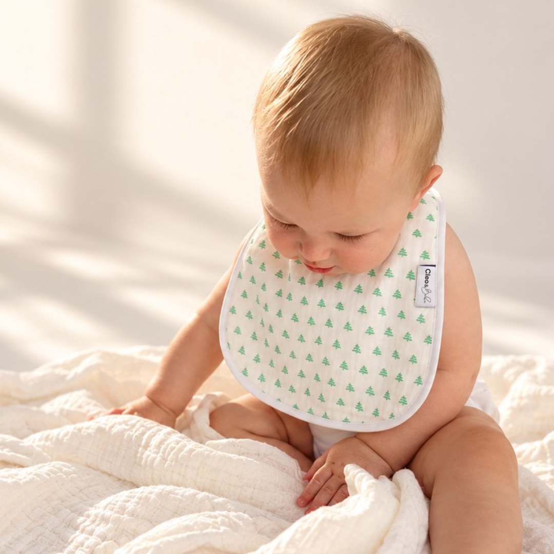A baby wearing a white bib with a green pine tree pattern sits on a white textured blanket in a brightly lit room.