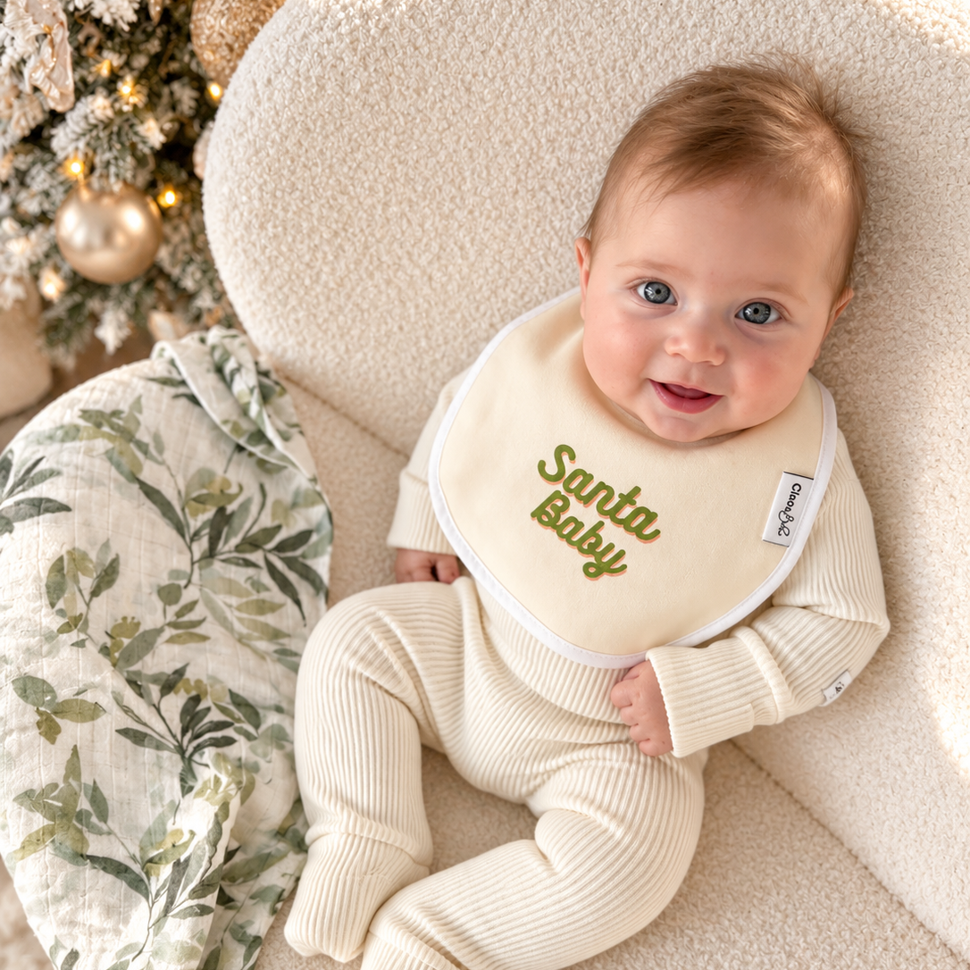 A baby wearing a cream ribbed footed one-piece and a matching cream bib with 'Santa Baby' embroidered in olive green sits on a textured cream chair in front of a decorated Christmas tree.