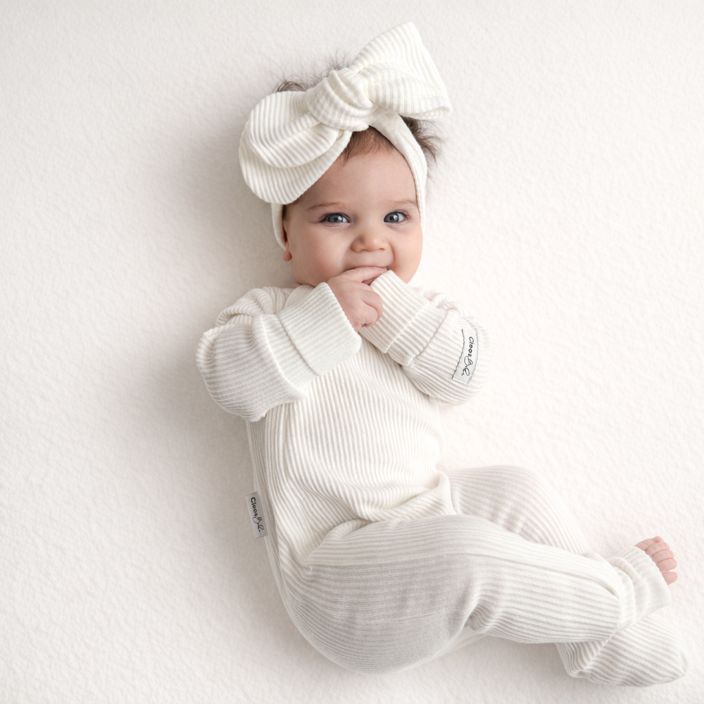A baby wearing a ribbed cream-colored footed romper and matching large bow headband, lying on a textured white surface.