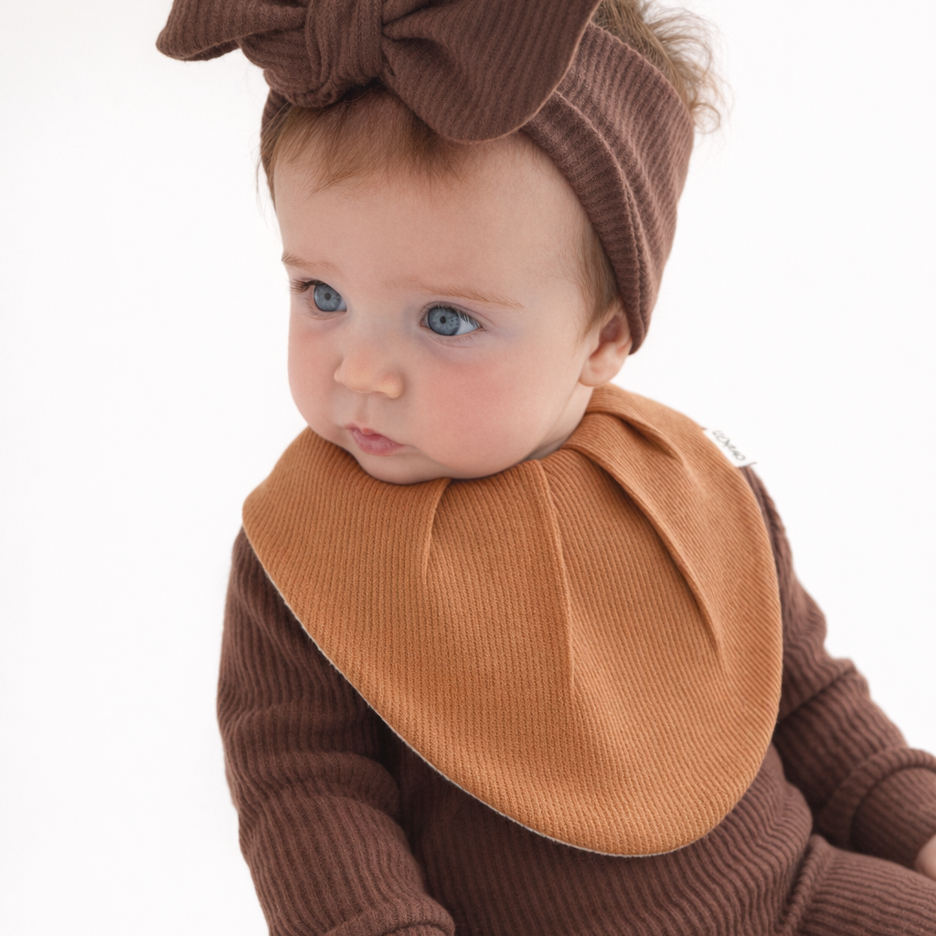 A baby wearing a brown ribbed corduroy footed jumpsuit, a matching brown bow headband, and a tan bib, sitting against a plain white background.