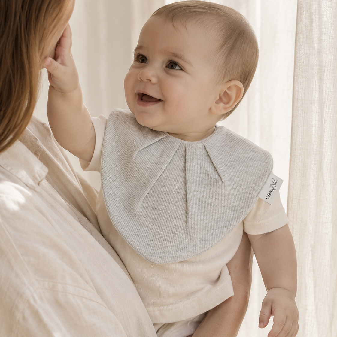 A baby wearing a light gray textured bib and cream outfit, held by a mother in a matching cream top against a soft, warm cream curtain background.