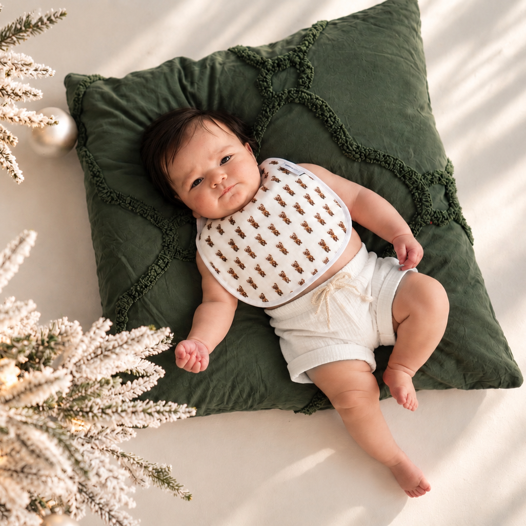 A baby lying on a dark green pillow next to a decorated Christmas tree, wearing a white bib with a brown reindeer pattern and white diaper cover.