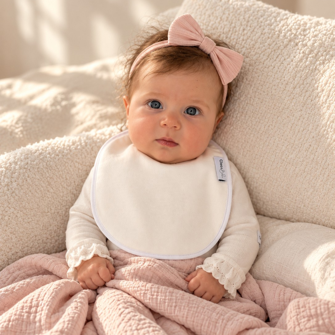 A baby wearing a white long-sleeve top, a white waterproof bib, and a pink bow headband, wrapped in a dusty pink textured blanket on a cream-colored textured chair.