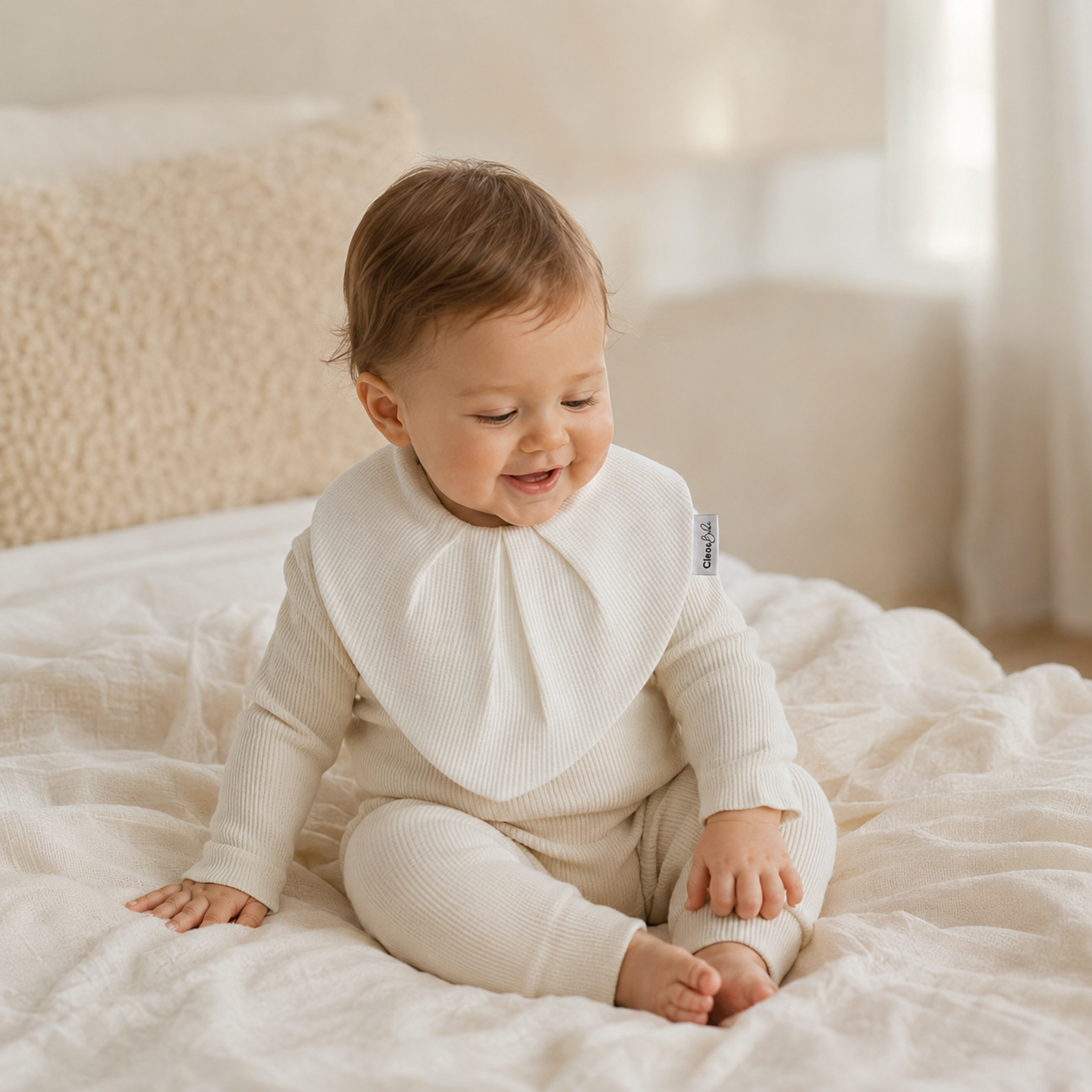A baby wearing a cream ribbed long-sleeve romper with a matching bib sits on a soft cream-colored bed. The brand tag on the side of the bib reads 'babi'.