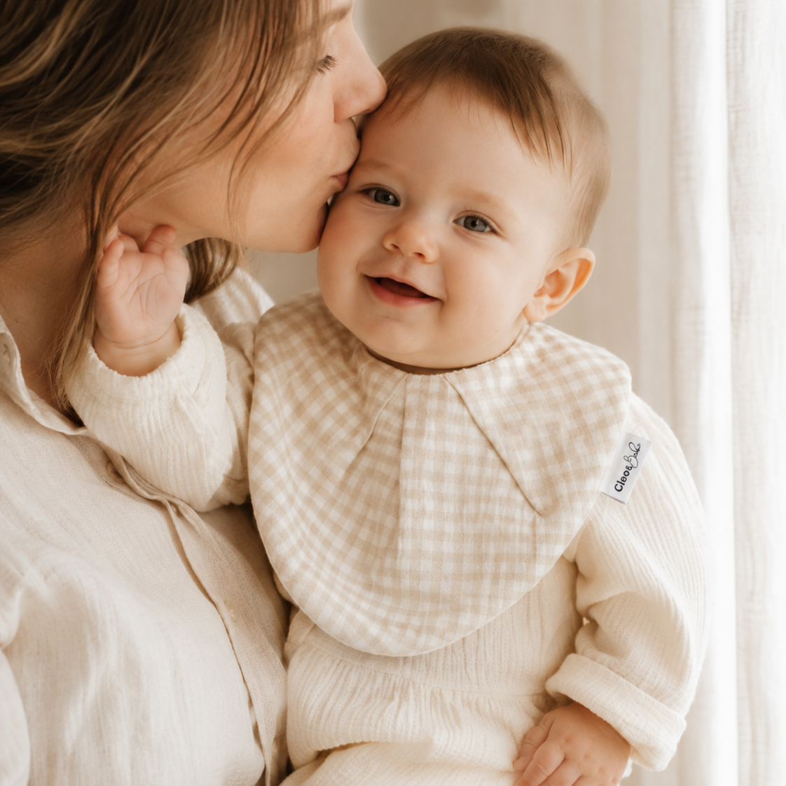 A baby wearing a cream ribbed long-sleeve romper and a beige and white gingham bib, being kissed on the cheek by a woman in a matching cream ribbed top, set against a soft, bright background with white curtains.