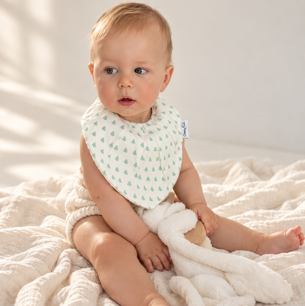 A baby sitting on a textured cream blanket, wearing a light green and white patterned bib and holding a white plush bunny toy.