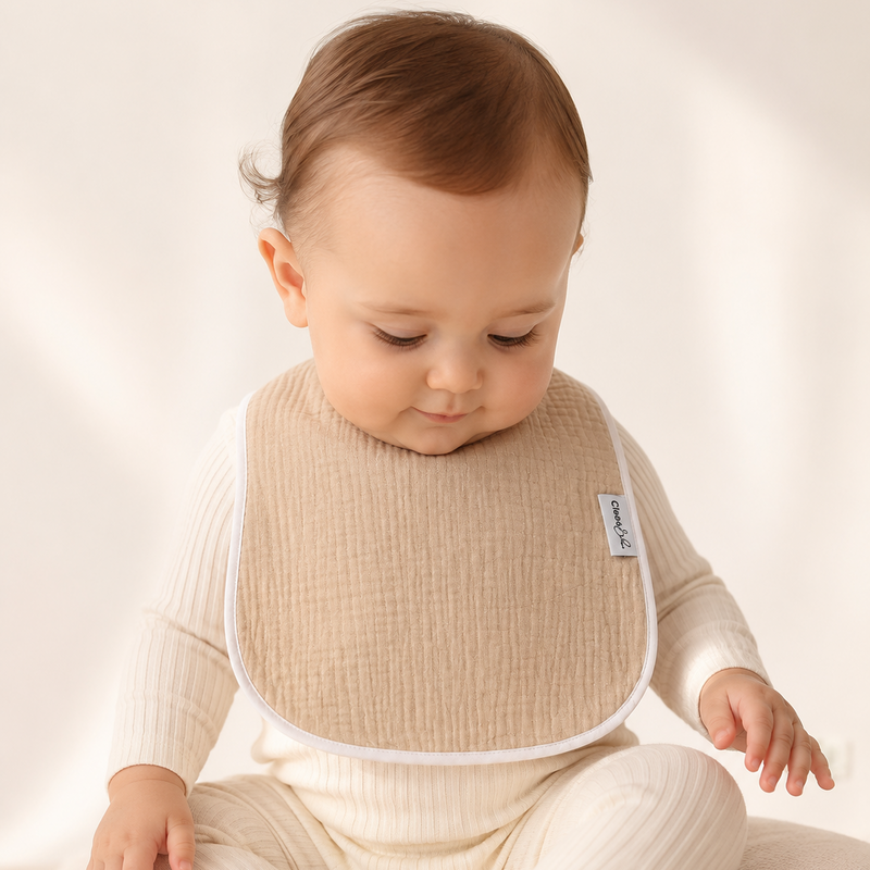 A baby wearing a cream ribbed long-sleeve outfit and a beige textured bib with white trim, sitting on a soft cream cushion against a warm off-white background.