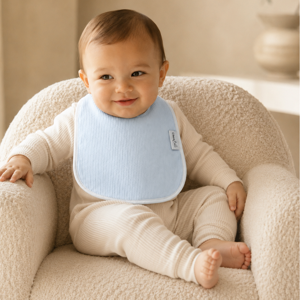 A baby wearing a light blue ribbed bib and cream ribbed one-piece outfit sits smiling in a textured cream armchair in a softly lit indoor setting.