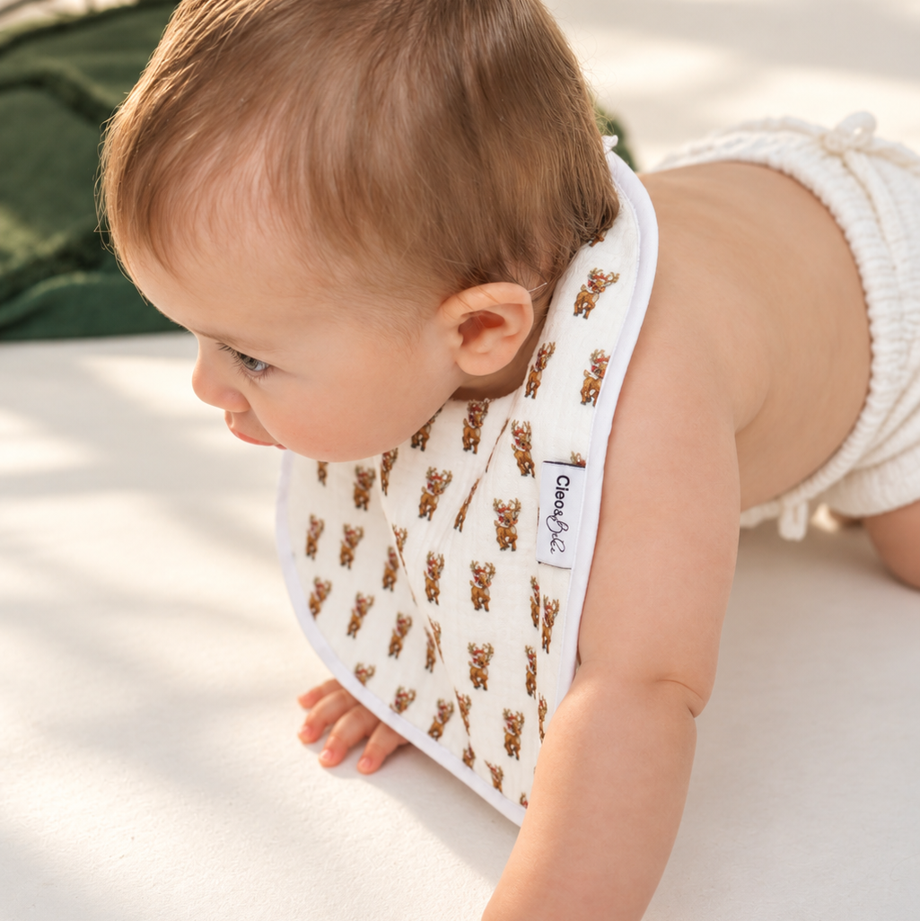 A baby crawling on a white floor wearing a white bib with a brown teddy bear pattern. A small white tag on the bib reads 'daisy's'.