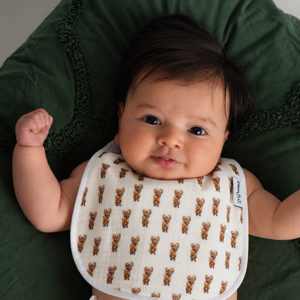 A baby lying on a dark green textured blanket, wearing a white bib with a repeating brown reindeer pattern.