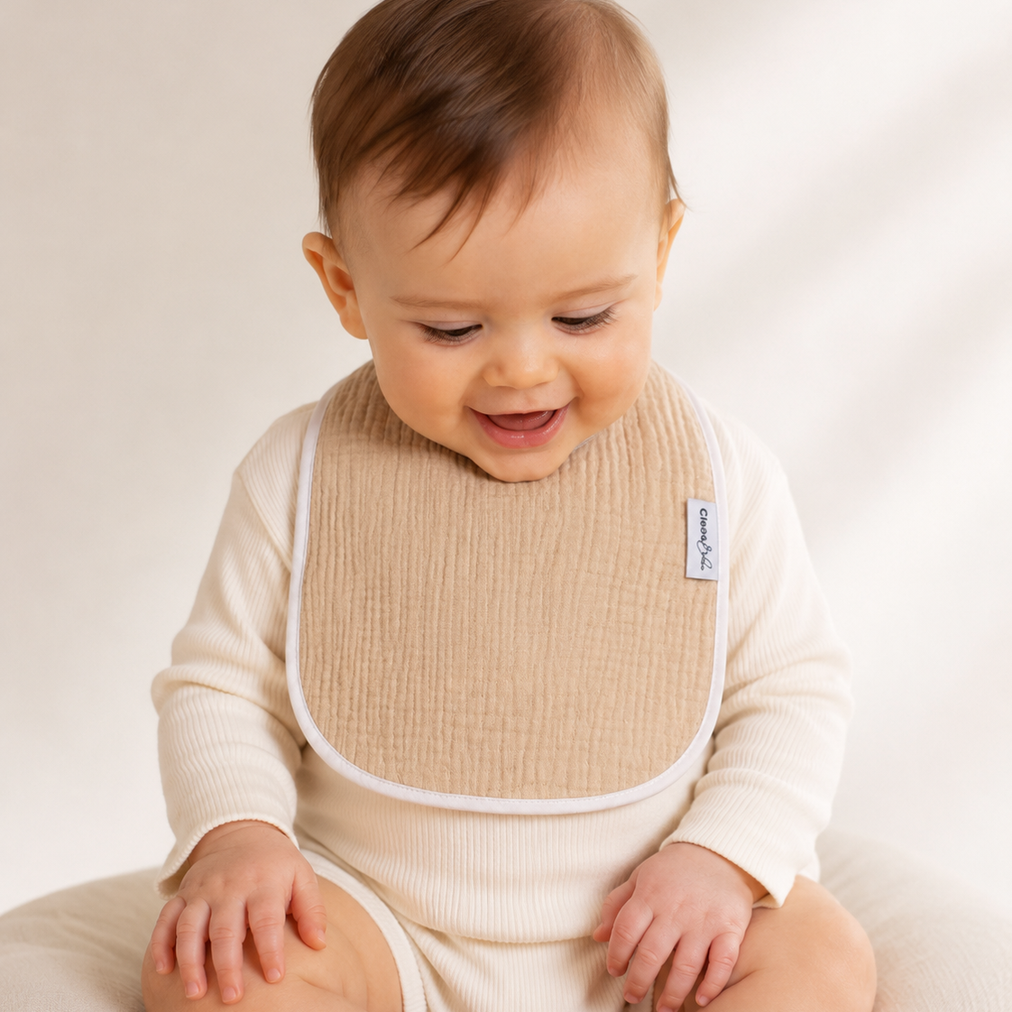 A baby wearing a cream ribbed long-sleeve bodysuit and a beige textured bib, sitting on a soft cream cushion against a warm off-white background.