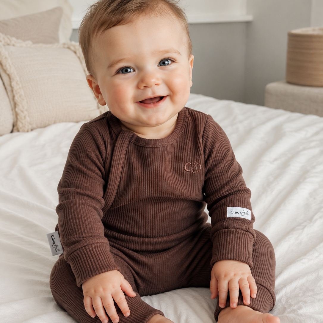 A smiling baby sitting on a white bed wearing a ribbed brown long-sleeve romper with a small embroidered logo on the chest and a brand patch on the sleeve.