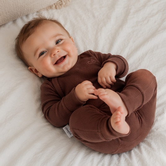 A four-panel collage showing a baby wearing a long-sleeve brown jumpsuit in different poses on a white bed. The baby is sitting, crawling, lying on their back, and standing while wearing the matching brown one-piece outfit.
