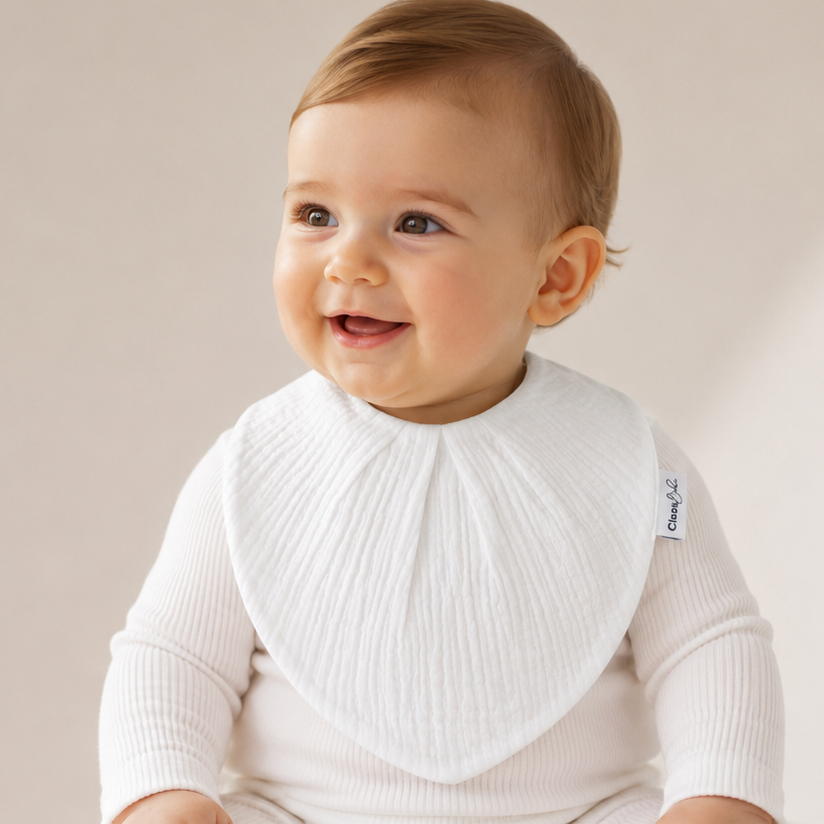 A baby sitting on a cushion wearing a white ribbed long-sleeve bodysuit and a white bib. The baby is looking to the side with a happy expression.