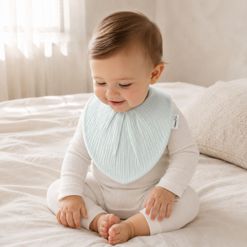 A baby wearing a light mint ribbed bib over a cream ribbed bodysuit, sitting on a white bed.