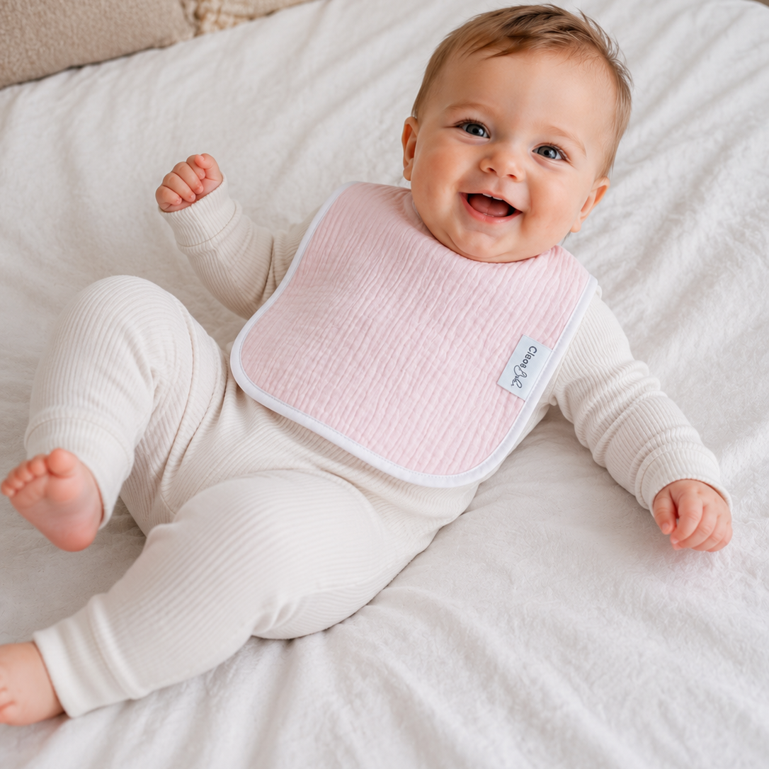 A smiling baby lying on a white bed, wearing a light pink textured bib with white trim and a small blue brand tag. The baby is also dressed in a cream ribbed long-sleeve footed onesie.