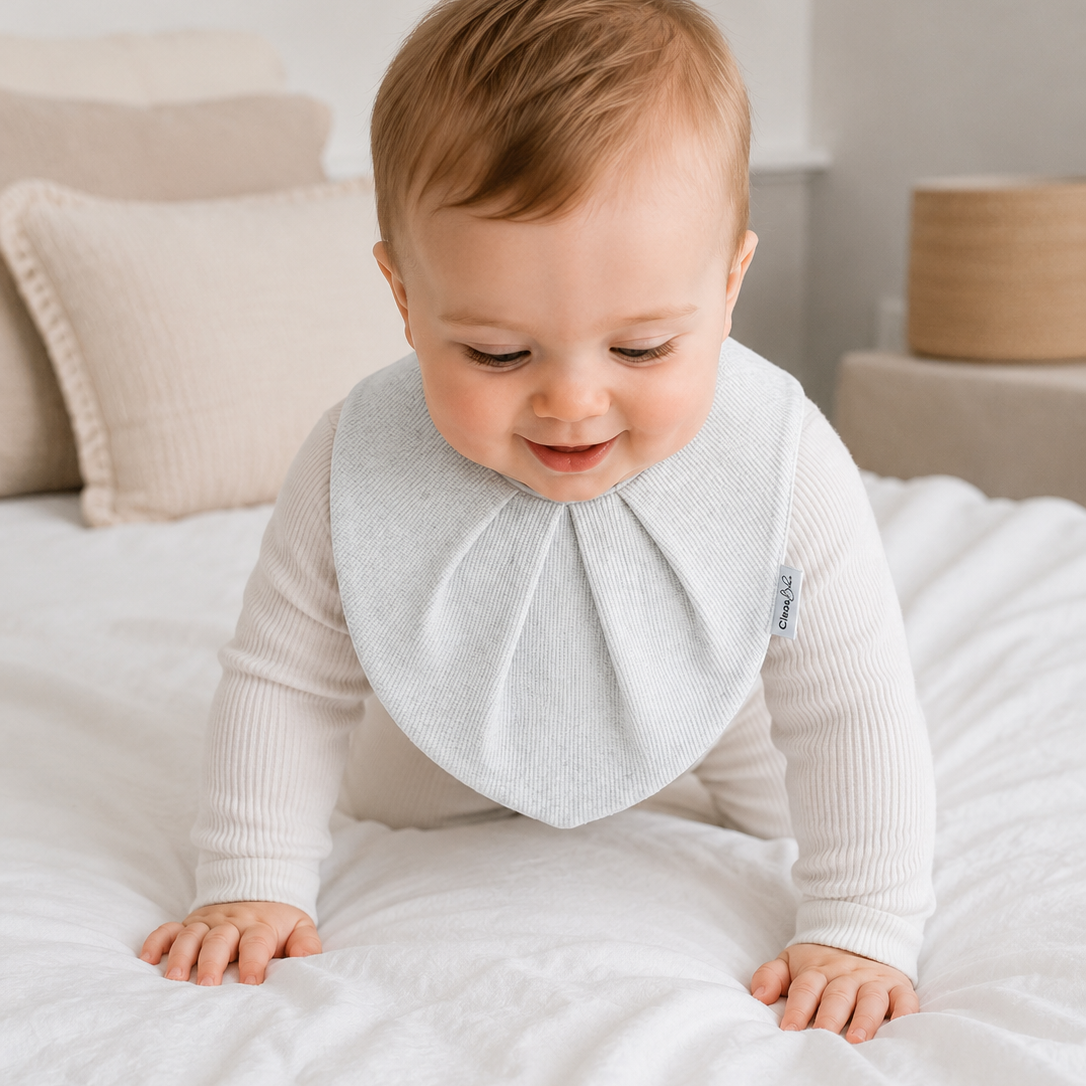A baby crawling on a white bed wearing a light gray bib with a long ribbed neck and a small blue tag on the side.