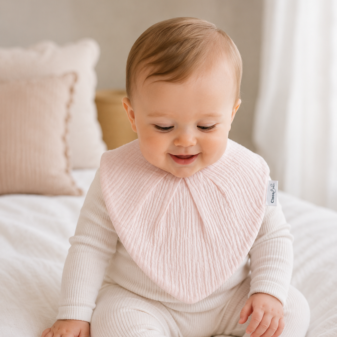 A baby wearing a pale pink ribbed bib over a cream ribbed one-piece outfit, sitting on a white bed in a softly lit room.