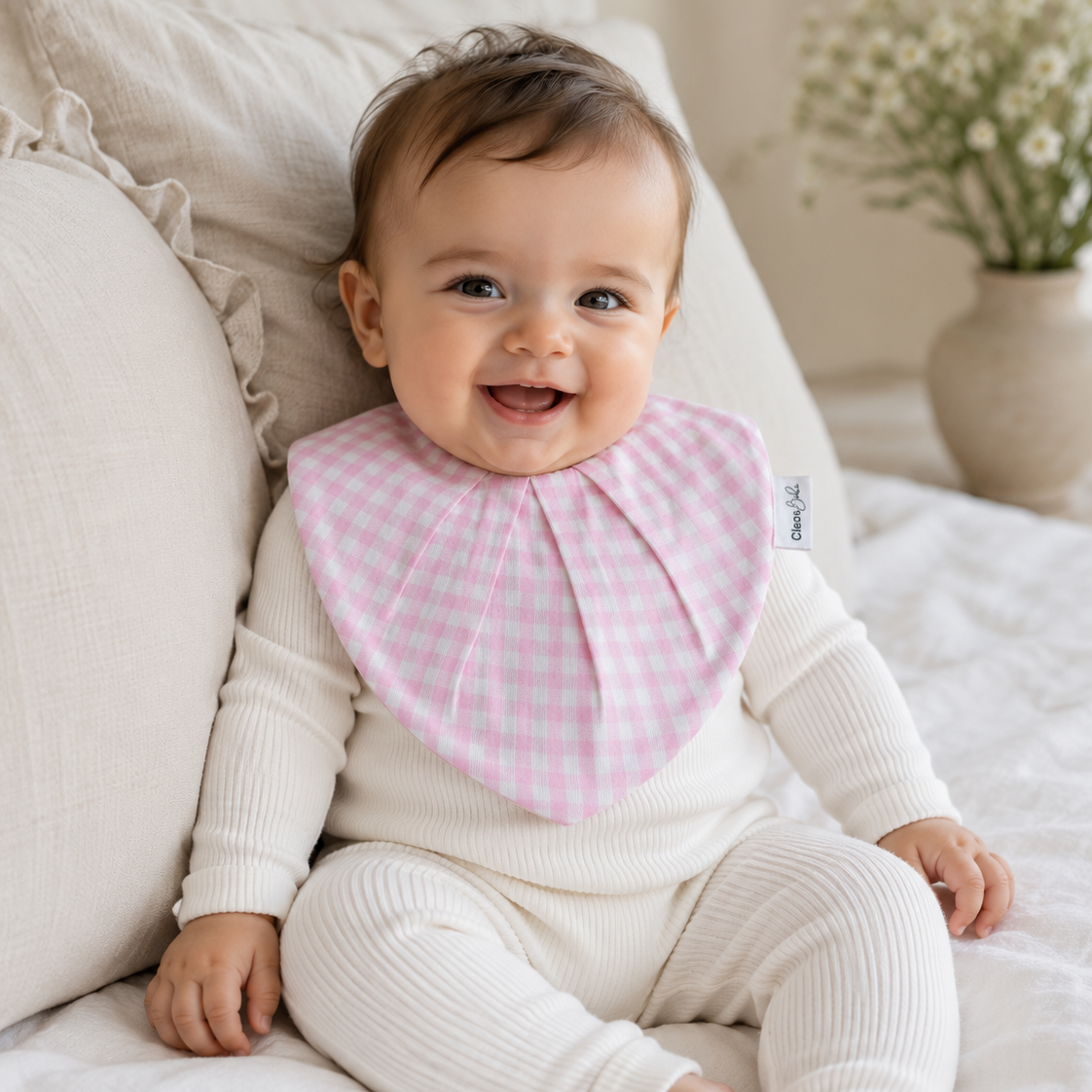 A smiling baby sitting on a white bed wearing a pink and white gingham bib over a cream ribbed romper, with pillows and a vase of flowers in the background.