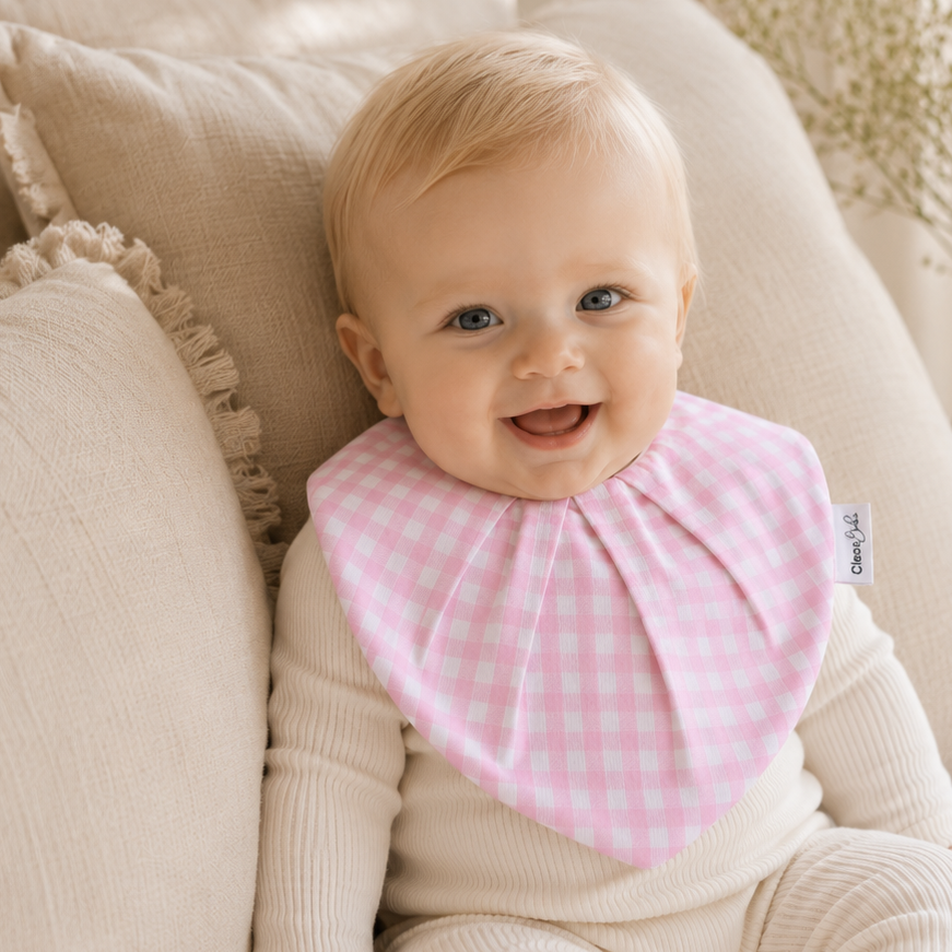 A baby wearing a pink and white gingham bib over a cream ribbed one-piece outfit, sitting on a bed with beige pillows and dried flowers in the background.