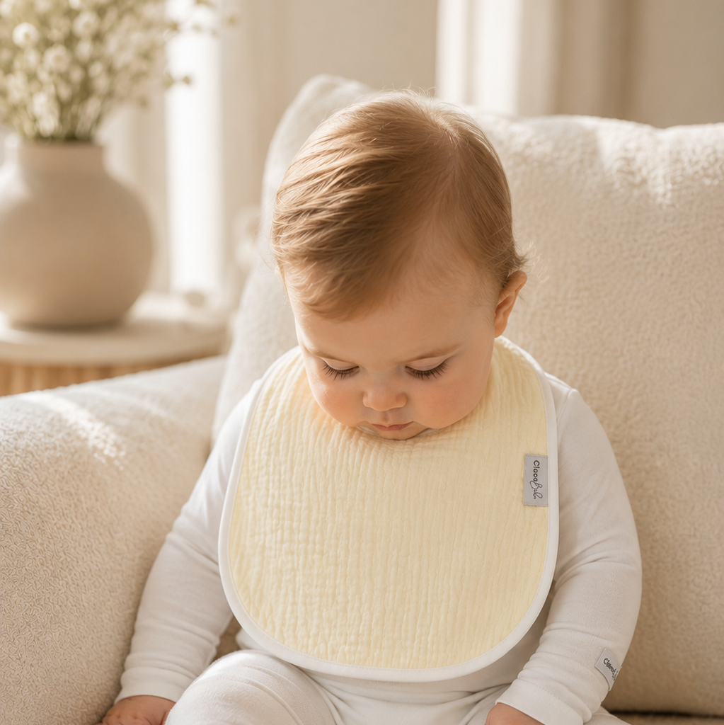 A baby wearing a cream-colored textured bib over a white long-sleeve footed romper, sitting on a soft beige couch in a brightly lit room.