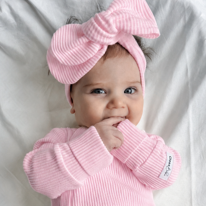 A baby wearing a pink ribbed one-piece jumpsuit and matching pink bow headband, lying on white fabric.