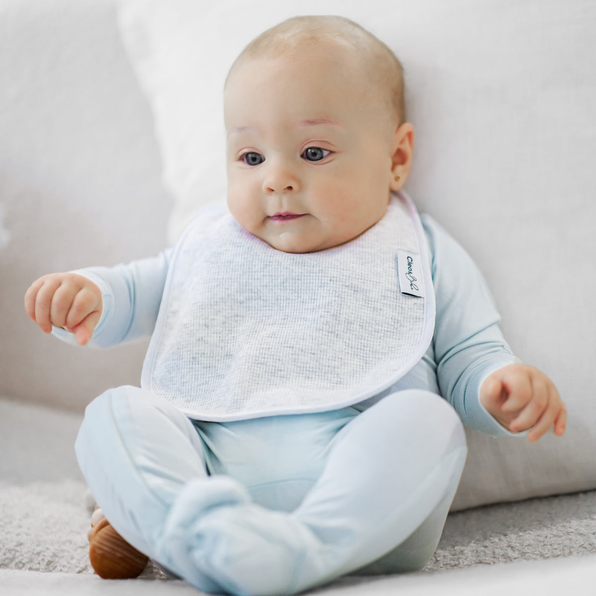 A baby wearing a light blue footed onesie and a marble grey waffle bib, sitting on a white sofa.