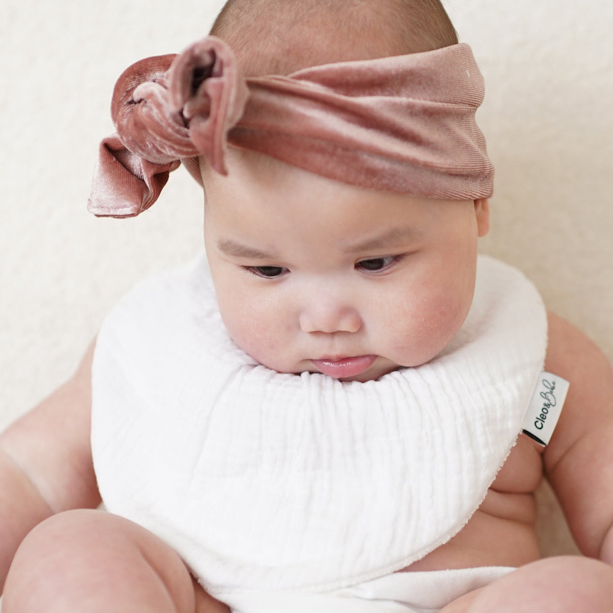 A baby sitting on a cream-colored upholstered chair, wearing a dusty rose velvet headband with a bow and a white bib.