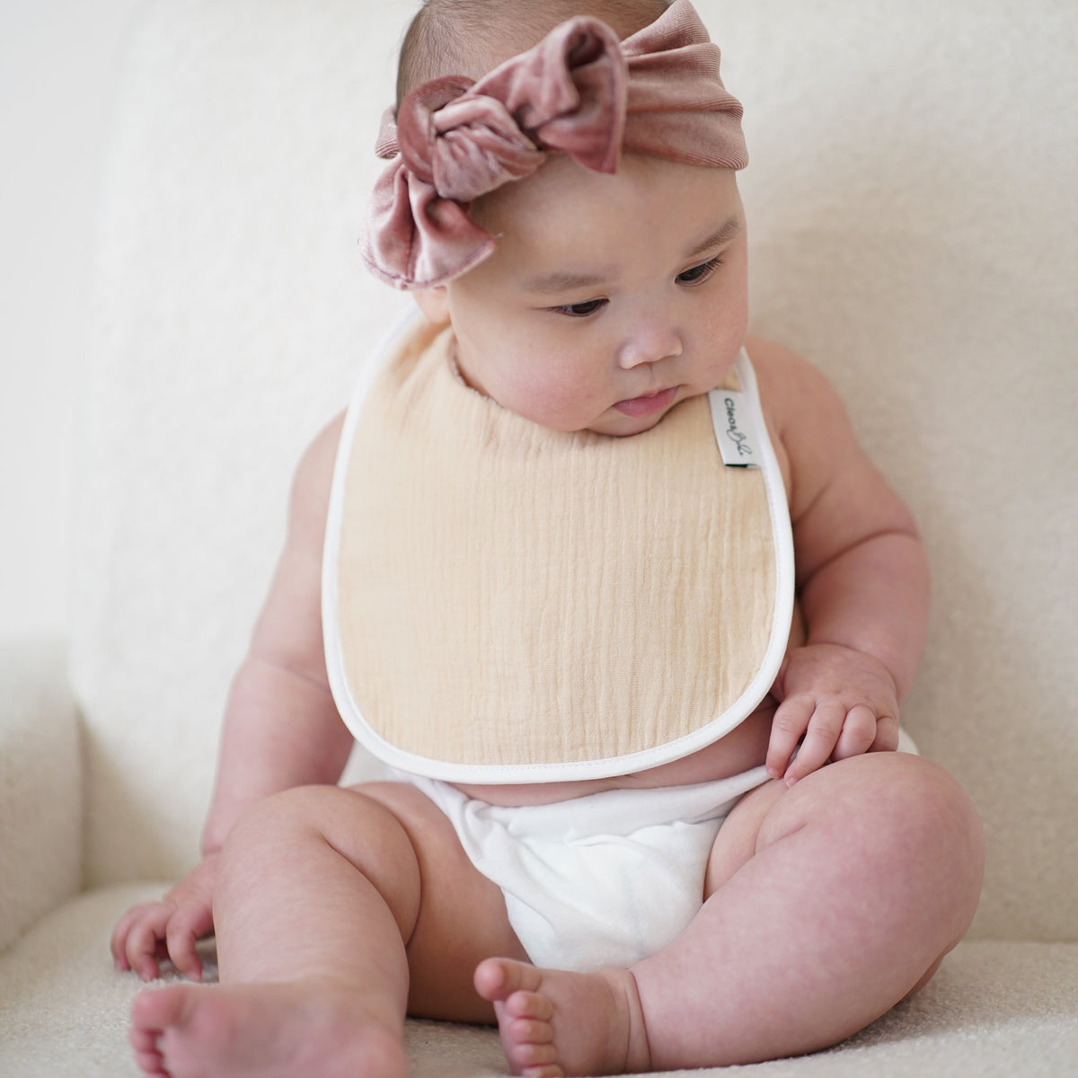 A baby sitting on a cream-colored armchair, wearing a dusty rose velvet bow headband and a muslin oat bib with a white border.