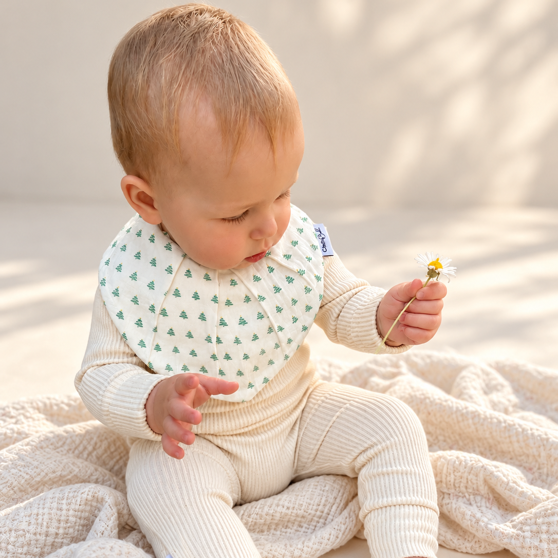 A baby sitting on a cream textured blanket, wearing a cream ribbed one-piece outfit and a white bib with a green pine tree pattern, holding a small yellow flower.