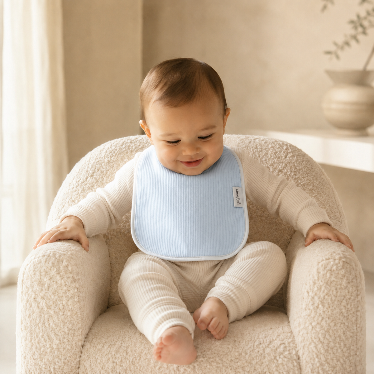 A baby wearing a light blue ribbed bib over cream ribbed one-piece clothing, sitting in a cream boucle armchair in a softly lit room.