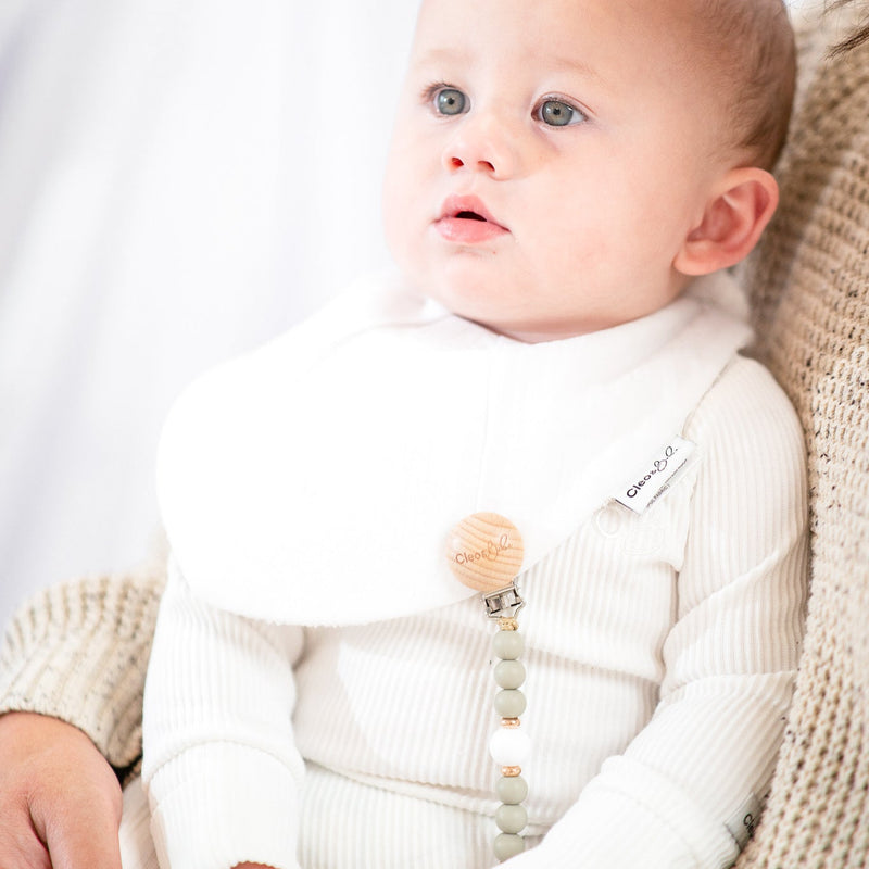 Baby wearing a white outfit with white bib and a pacifier clip, sitting on a person's lap 