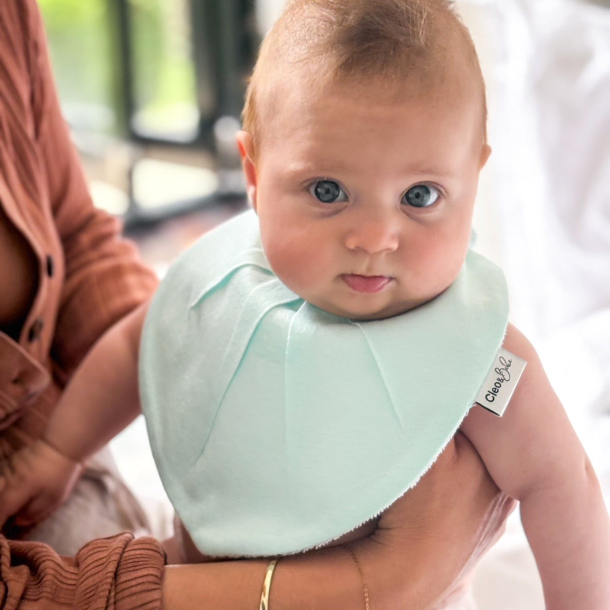 Baby wearing a light green bib with a brand label, held by an adult in a casual setting.