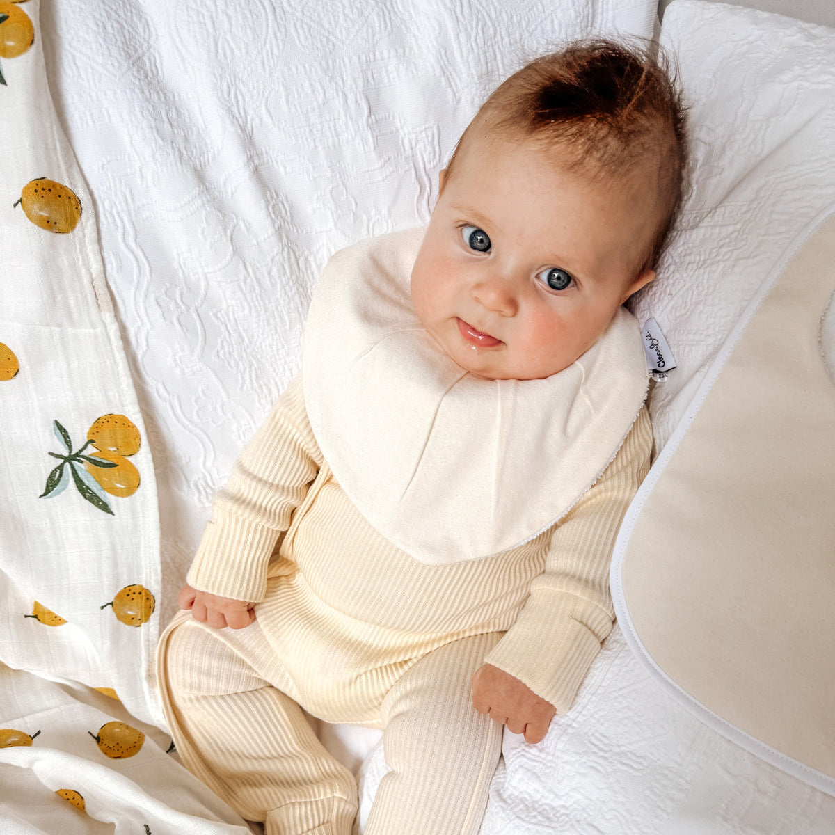 Baby in a cream-colored outfit lying on a white blanket with a lemon-patterned blanket.