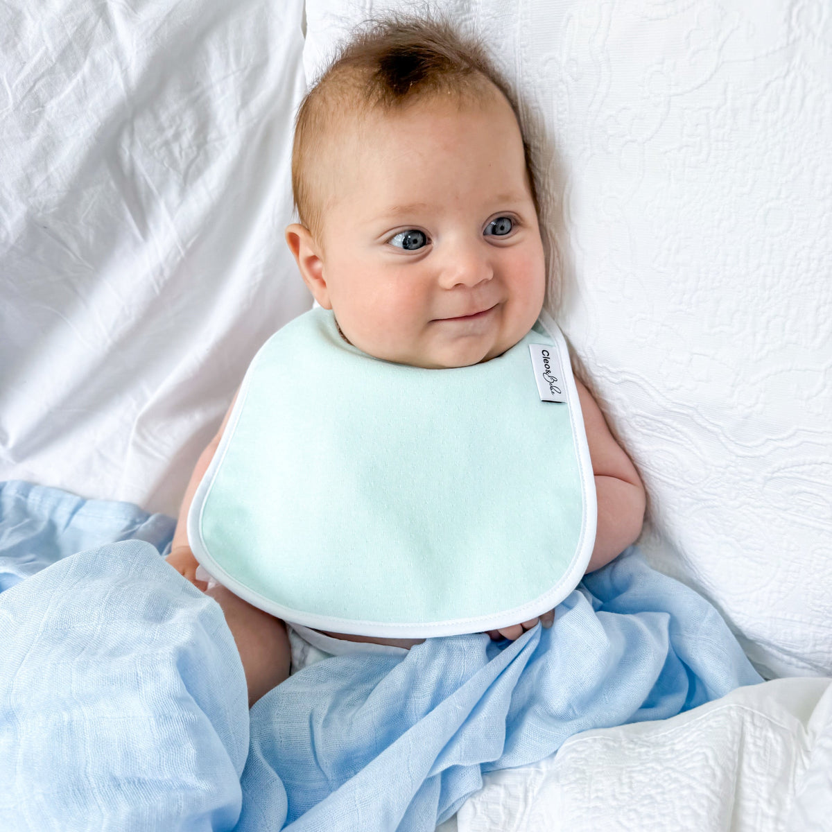 Baby wearing a light blue bib and surrounded by white and light blue blankets