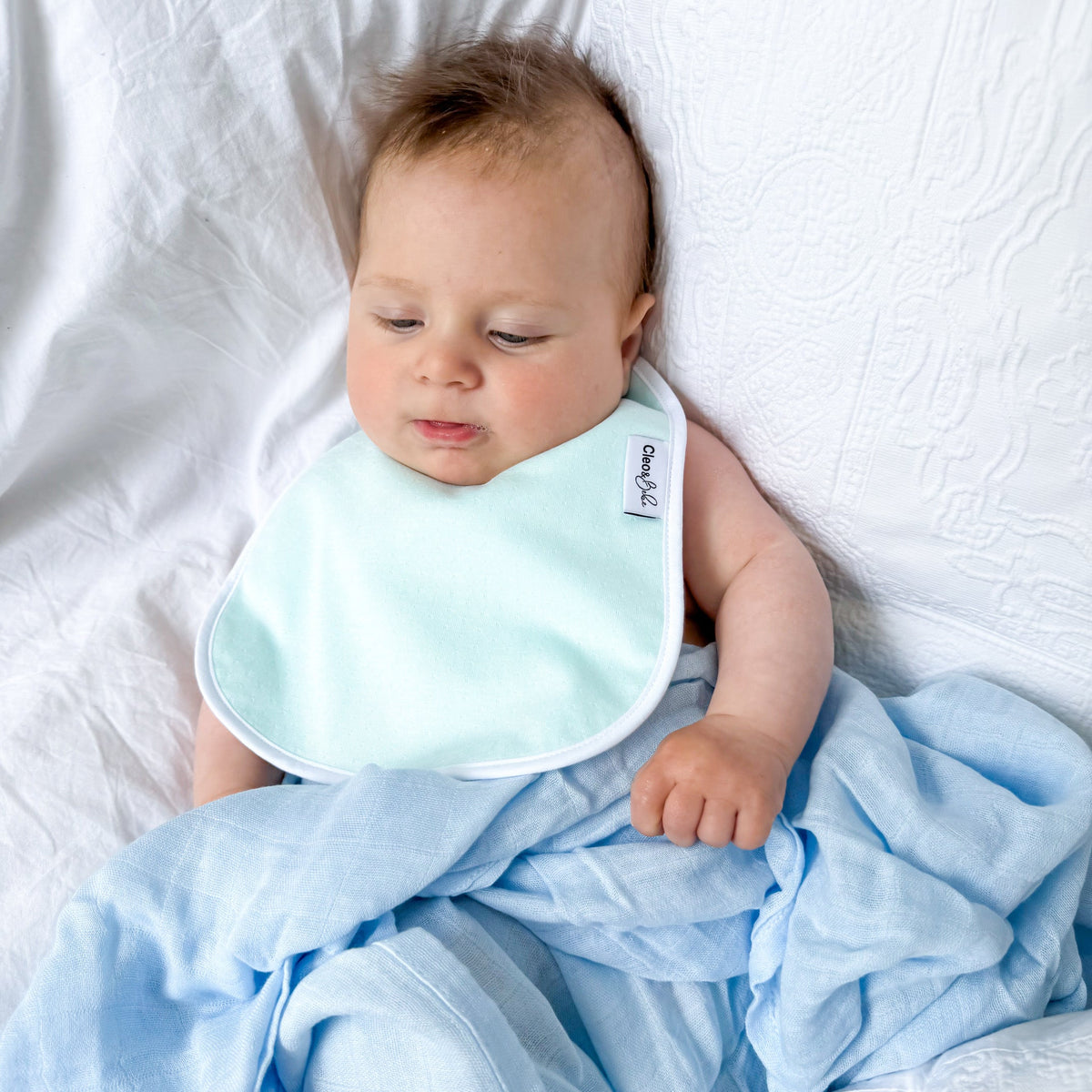 Baby lying on a white blanket with a light blue bib and blanket