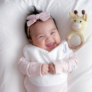 Baby lying on a white blanket with a pink bow and toy, wearing a white bib.