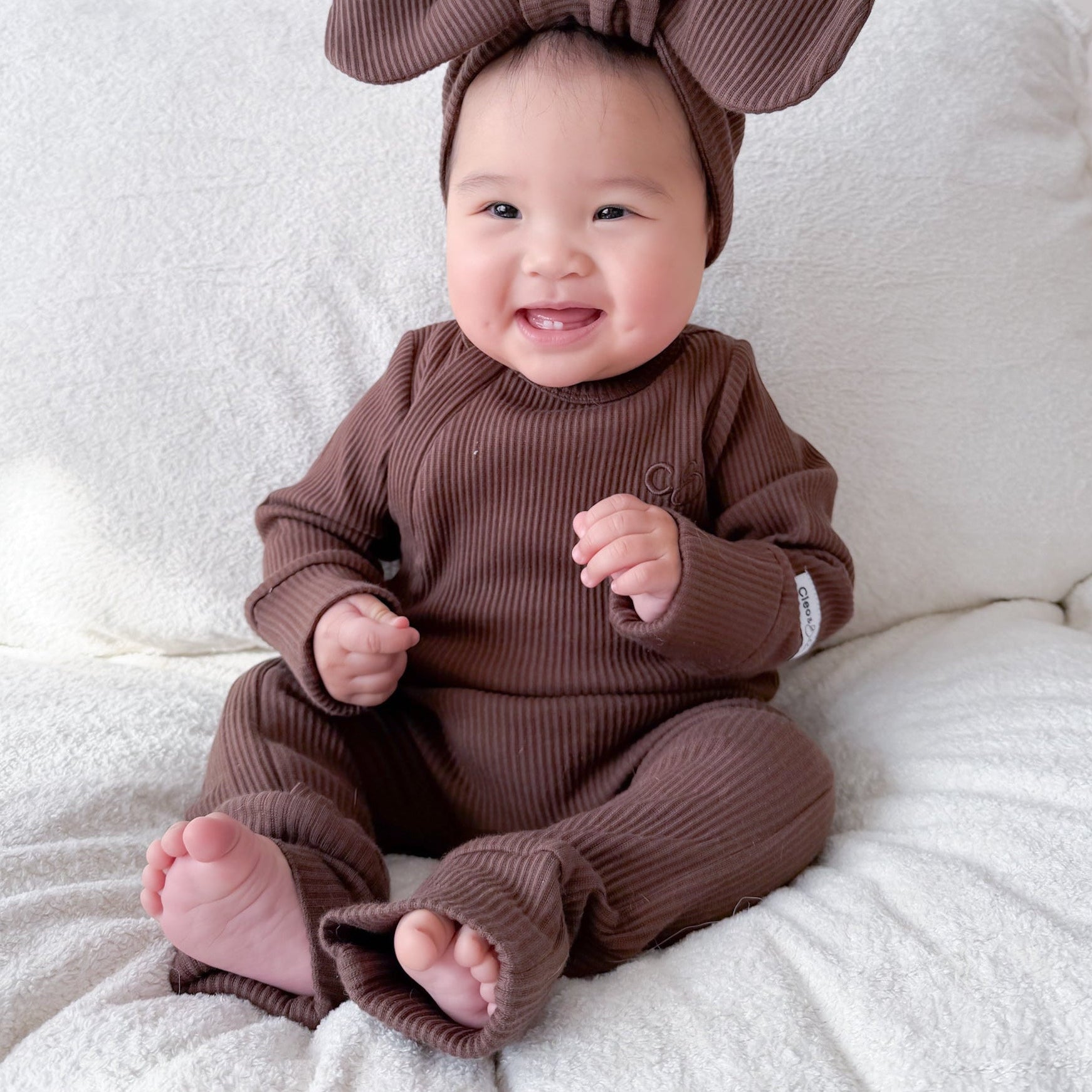 A smiling baby sitting on a white textured blanket, wearing a matching brown ribbed one-piece outfit with a large bow headband.