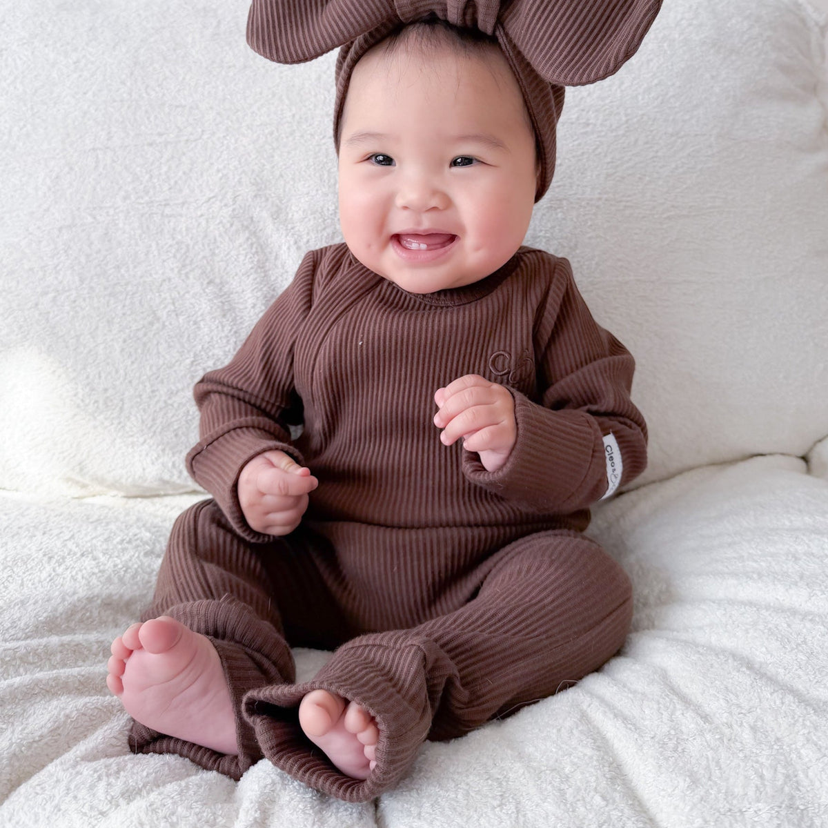 A smiling baby sitting on a white textured blanket, wearing a matching brown ribbed one-piece outfit with a large bow headband.
