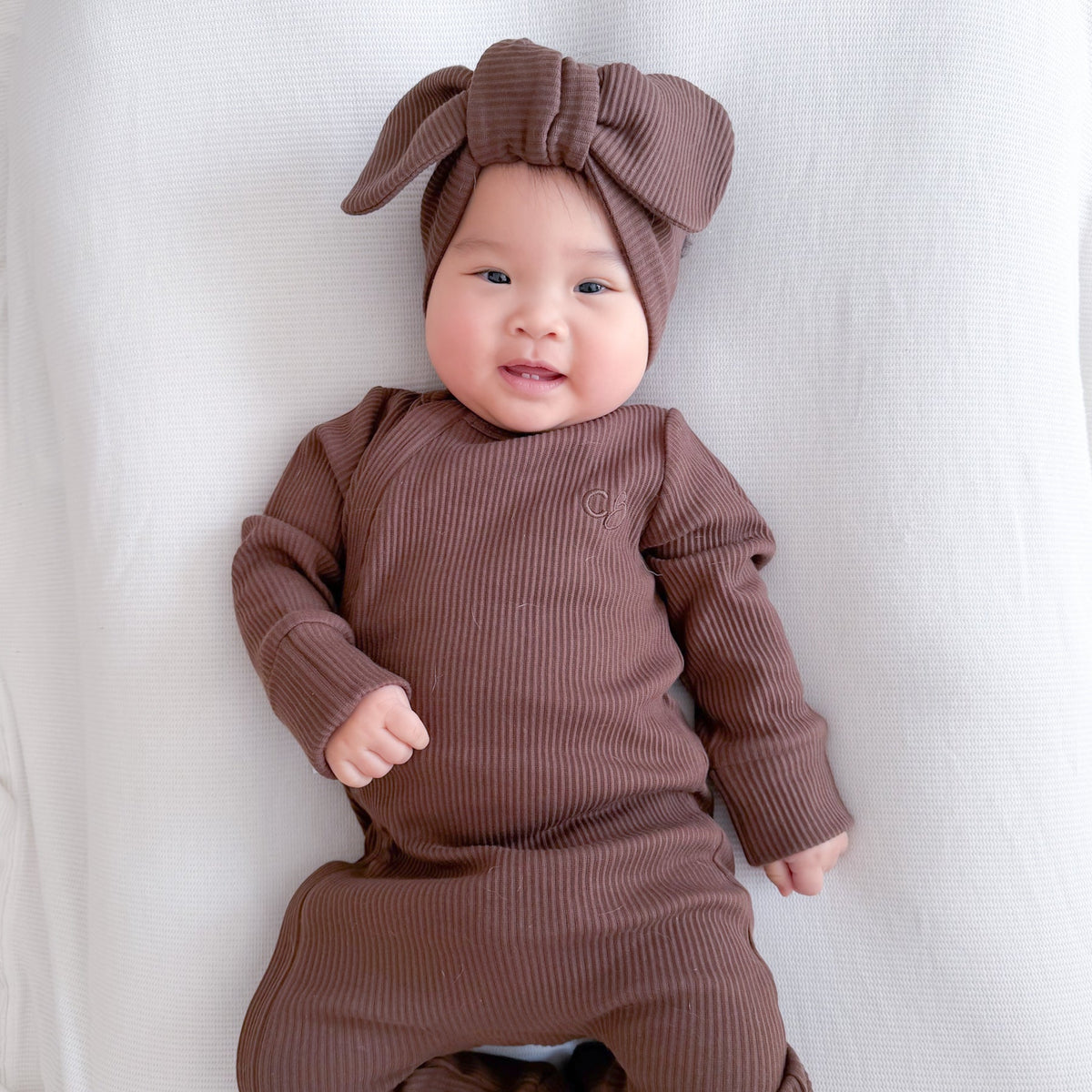 A baby lying on a white bassinet wearing a ribbed brown long-sleeve footed romper and matching bow headband.