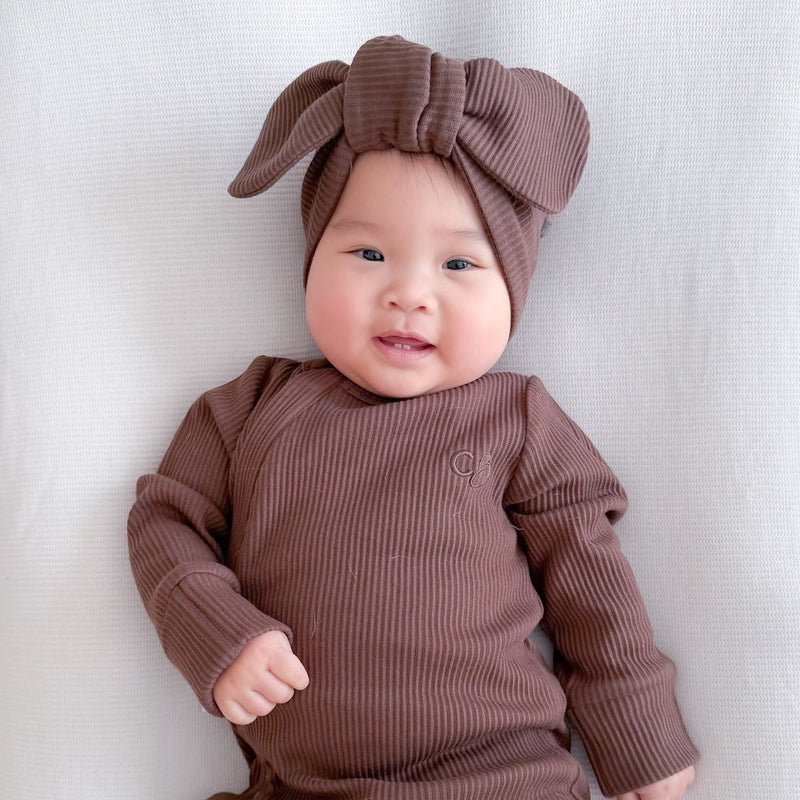 A baby lying on a white bassinet wearing a ribbed brown long-sleeve footed romper and matching bow headband.