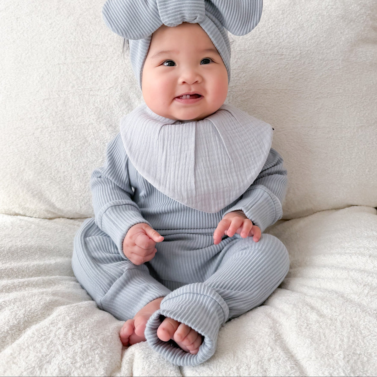 A baby sitting on a cream-colored textured sofa, wearing a light blue-gray ribbed knit footed one-piece outfit with a matching bow headband and a large bib.