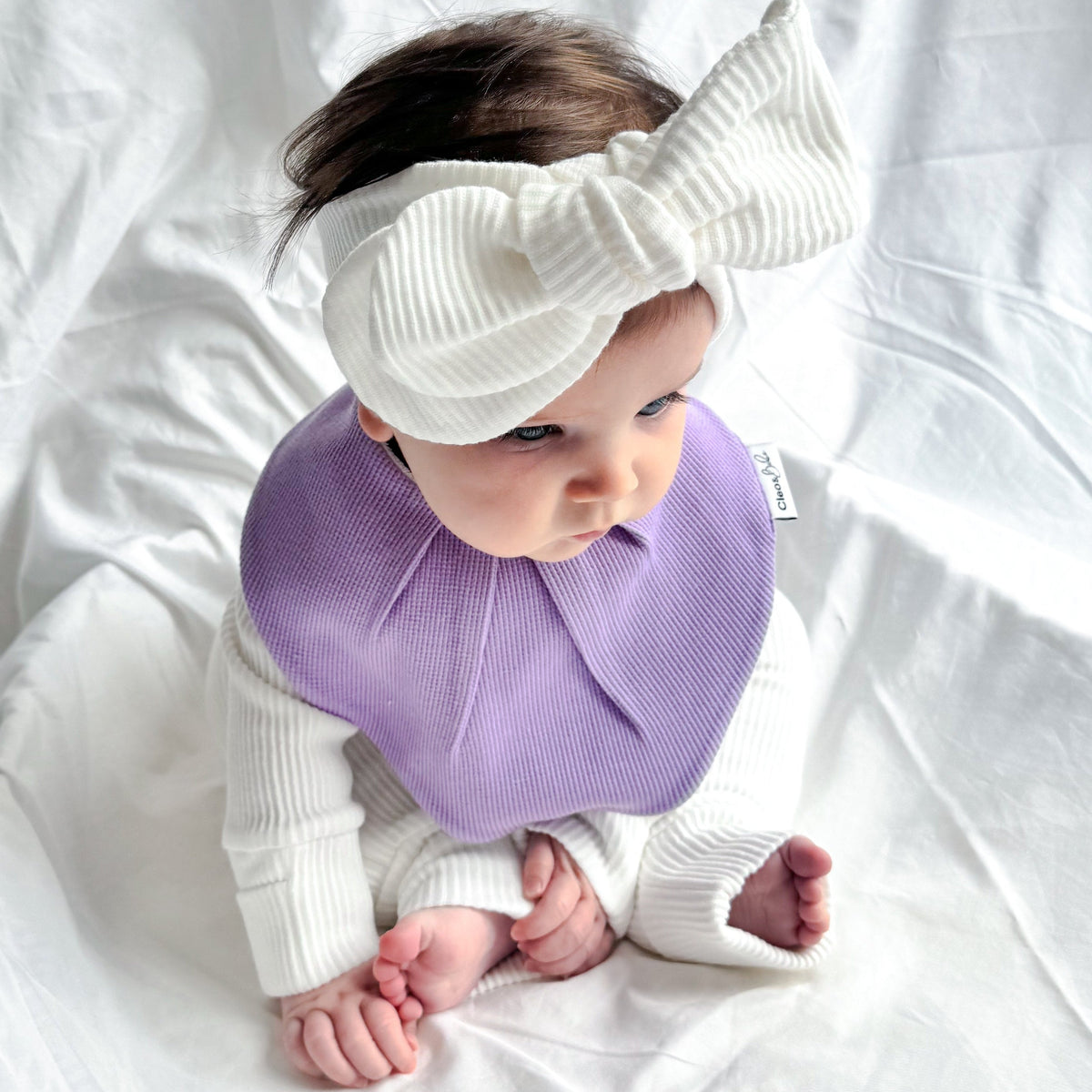 A baby wearing a white ribbed long-sleeve outfit, a lavender bib, and a large white bow headband, sitting on a white fabric backdrop.